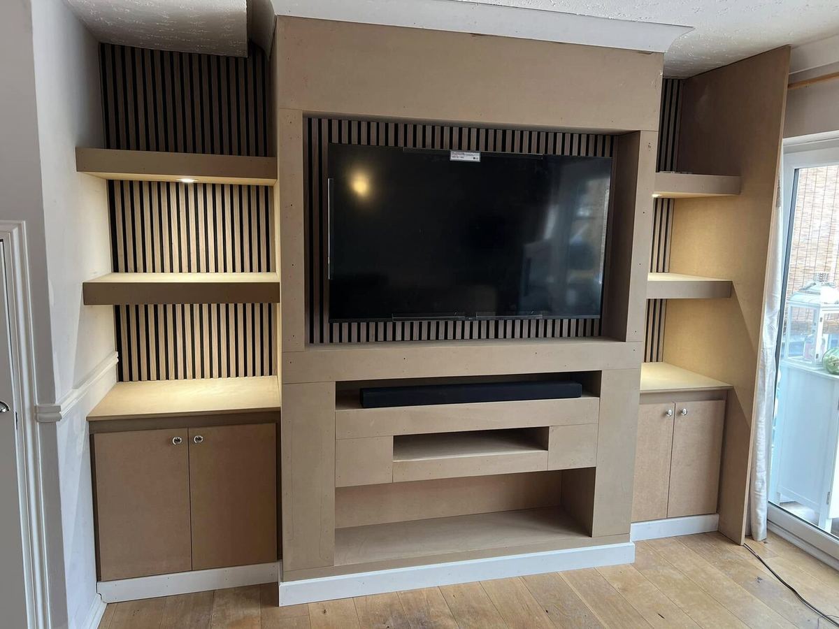 Bespoke media wall built into an alcove in bare MDF, with slatted timber panelling behind the television, LED-lit floating shelves and base cupboards